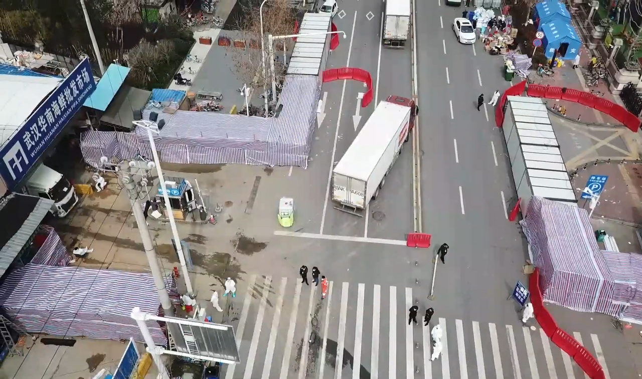 Elevated view of a city street junction: a market frontage under striped tarpaulins is sealed behind red barriers, a lorry stands at the kerb, and several figures in white protective suits work near a marked crossing.