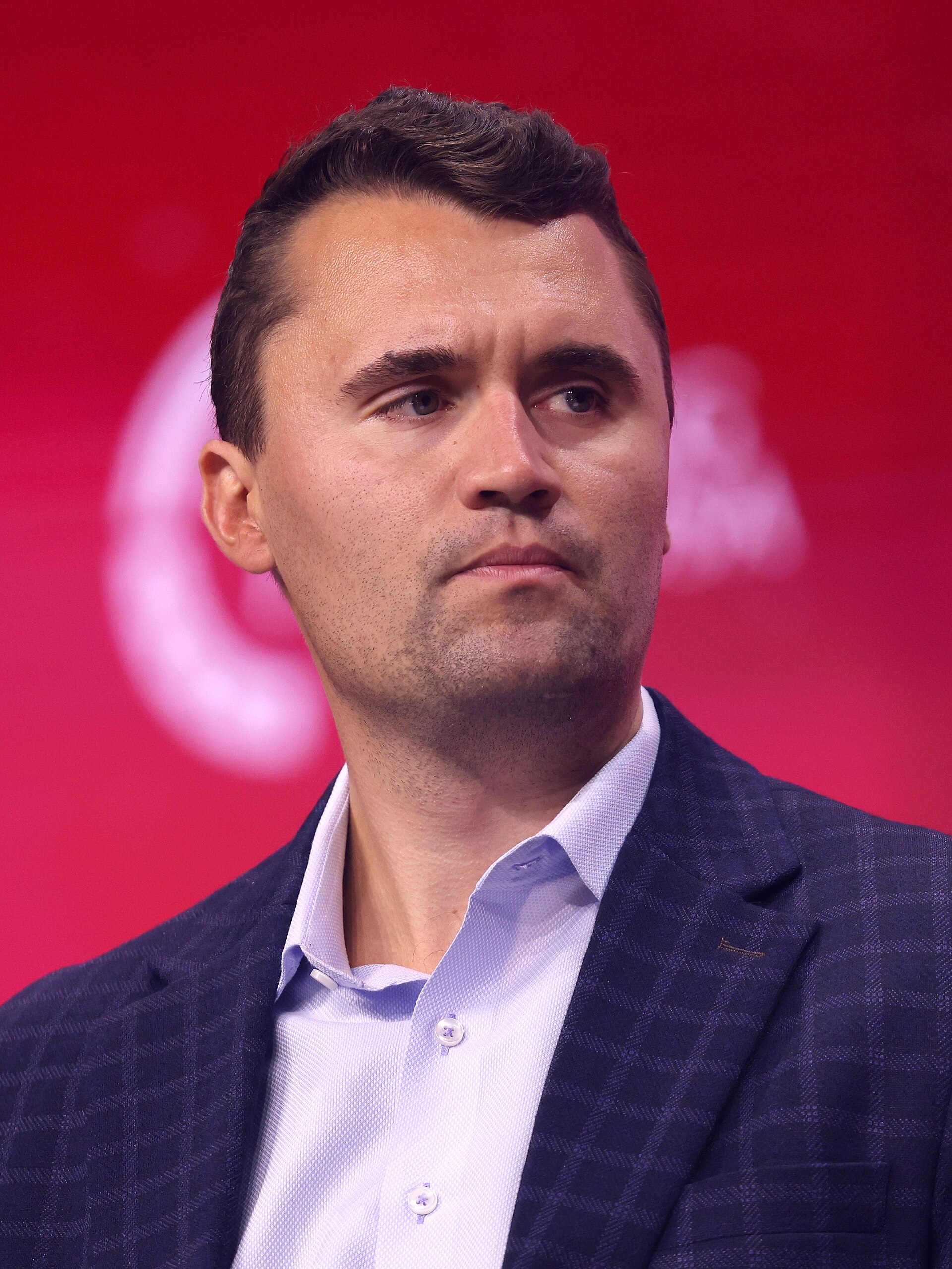 Charlie Kirk, in a dark checked blazer and open-collared shirt, looking off-camera against a red backdrop at a public event.