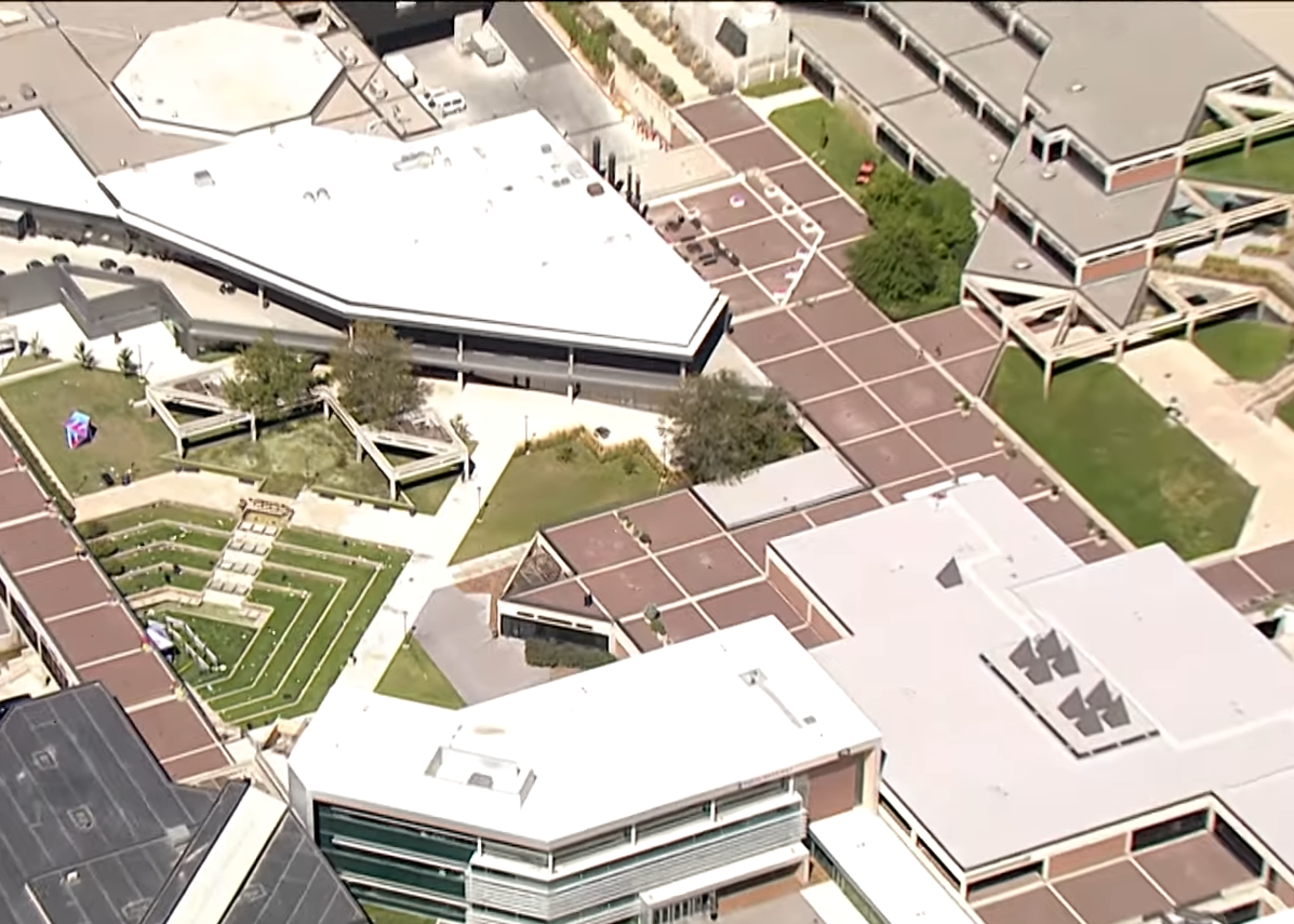 Aerial view of a university campus: a terraced courtyard with white event canopies, lawns, and walkways between large flat-roofed buildings.