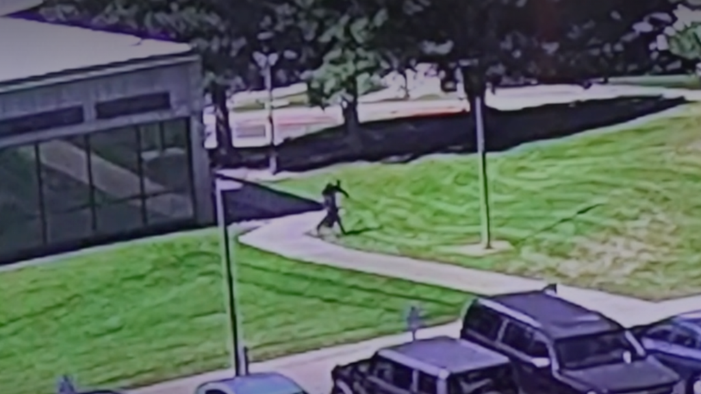 Grainy elevated surveillance frame: a distant, unidentifiable figure runs along a curved path across a campus lawn beside a glass-walled building, with parked cars in the foreground.